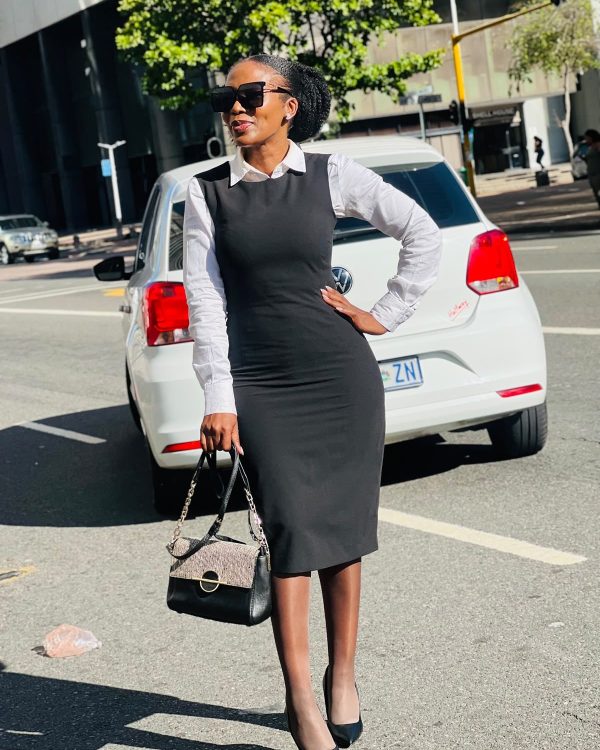 Woman in a black pinafore midi dress over a white shirt holding a handbag beside a white car